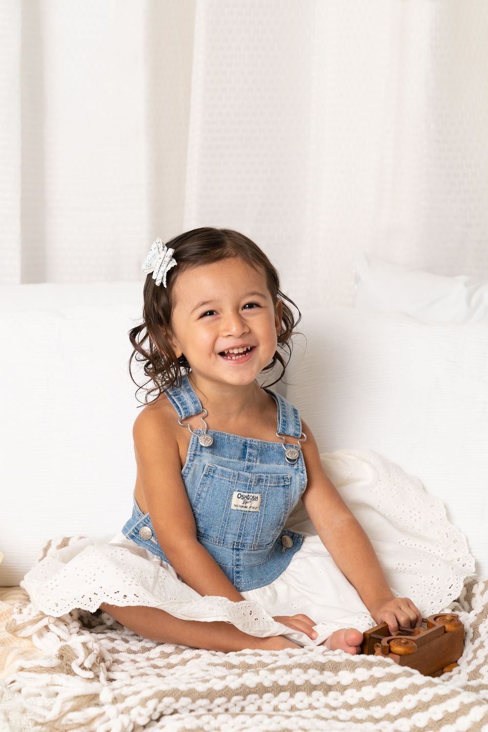 Smiling toddler girl in denim overalls with white flower hair clip sitting on couch with knit blanket.