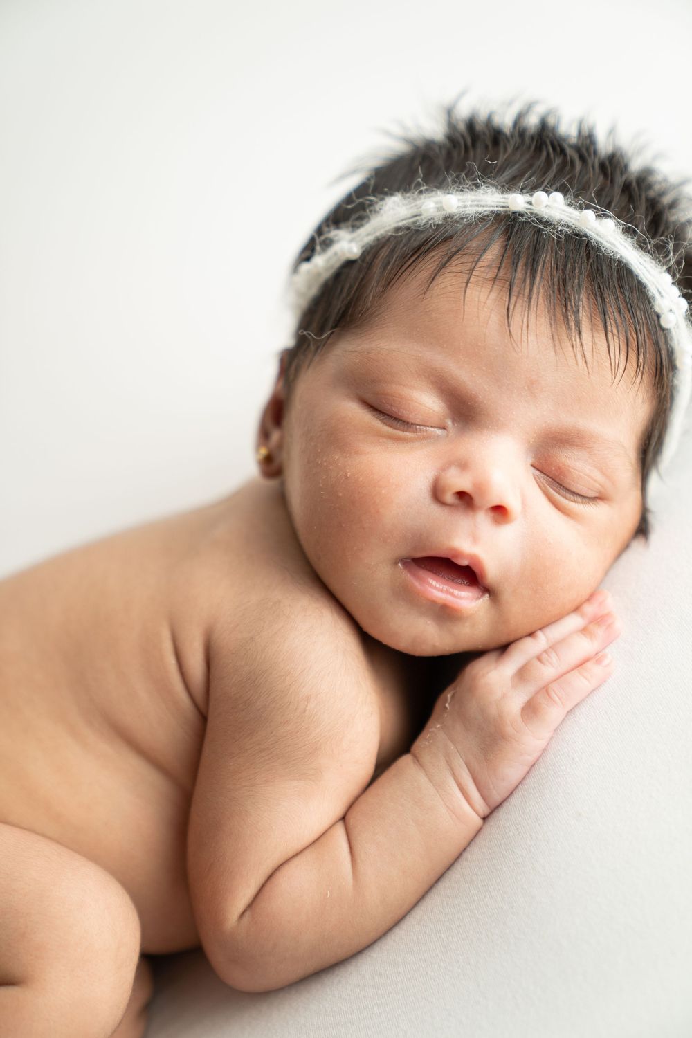 Sleeping newborn baby girl with dark hair wearing a white floral headband, lying on a soft white blanket.