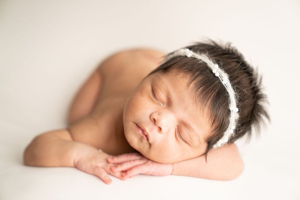 Sleeping newborn baby girl with dark hair wearing a white floral headband, resting peacefully on a white background.
