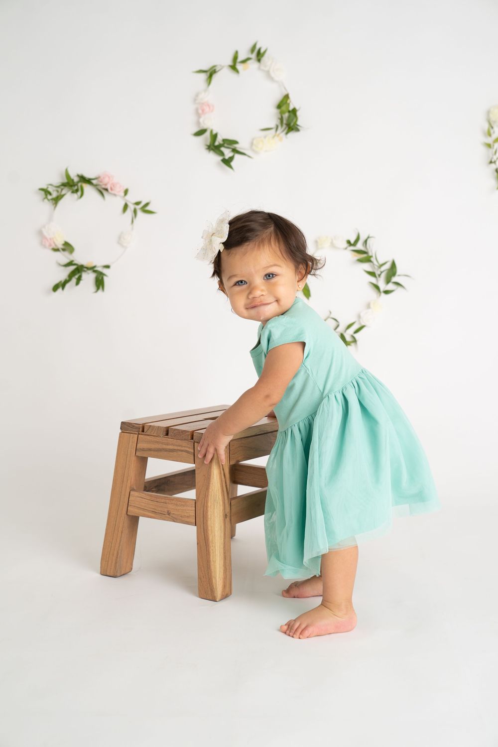 Happy toddler girl in teal dress stands beside wooden stool in white studio with floral wreaths backdrop.