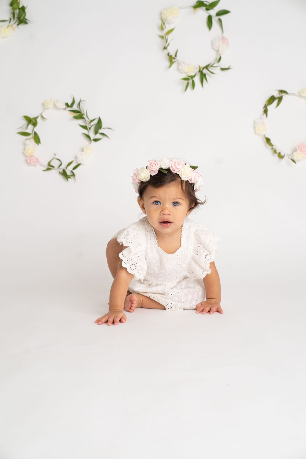 Baby girl in white lace romper and floral crown crawls on white floor with heart-shaped floral wreaths in background.