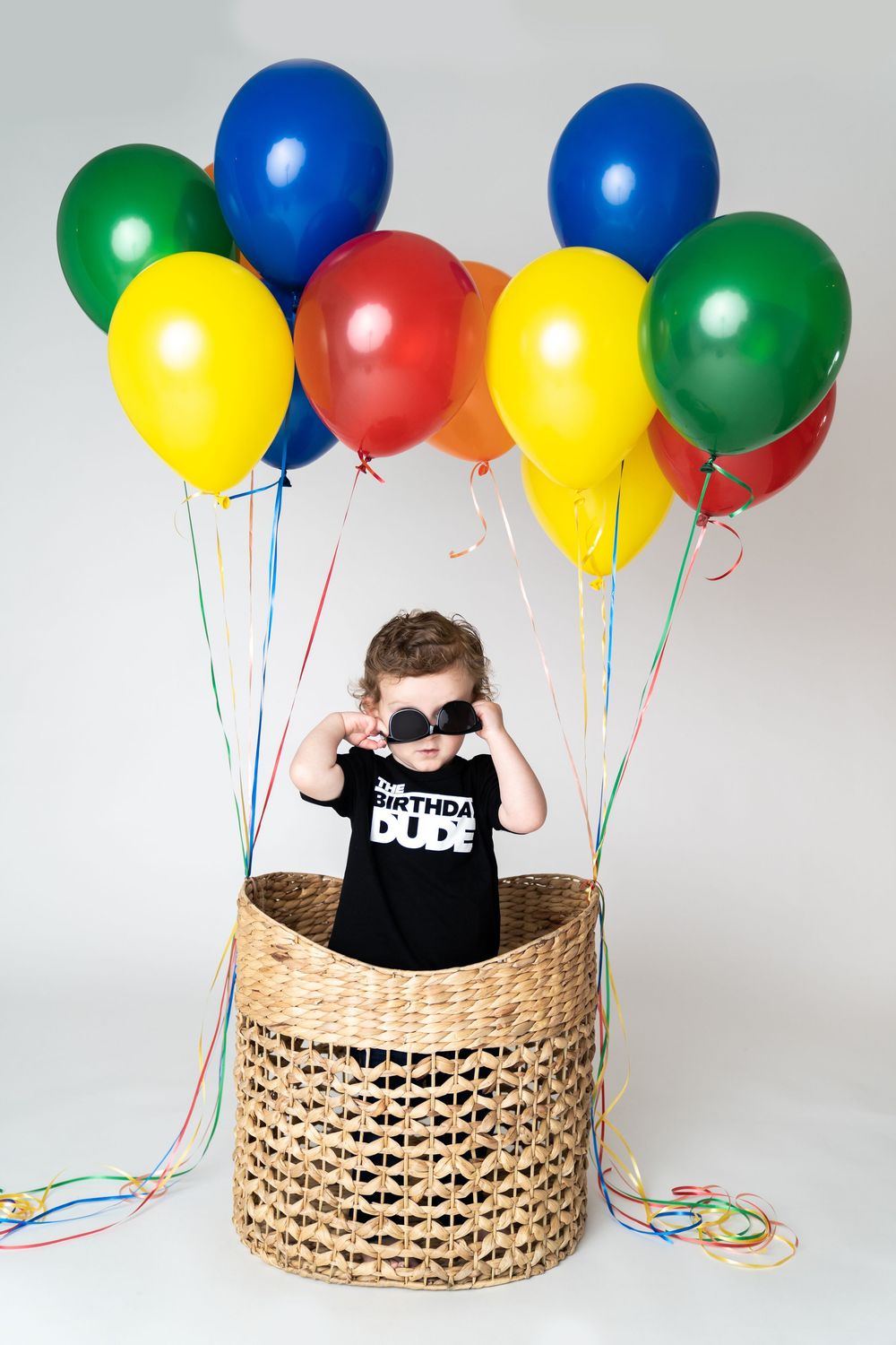 Toddler in a black Birthday Dude shirt wearing sunglasses sits in a wicker basket surrounded by colorful balloons.