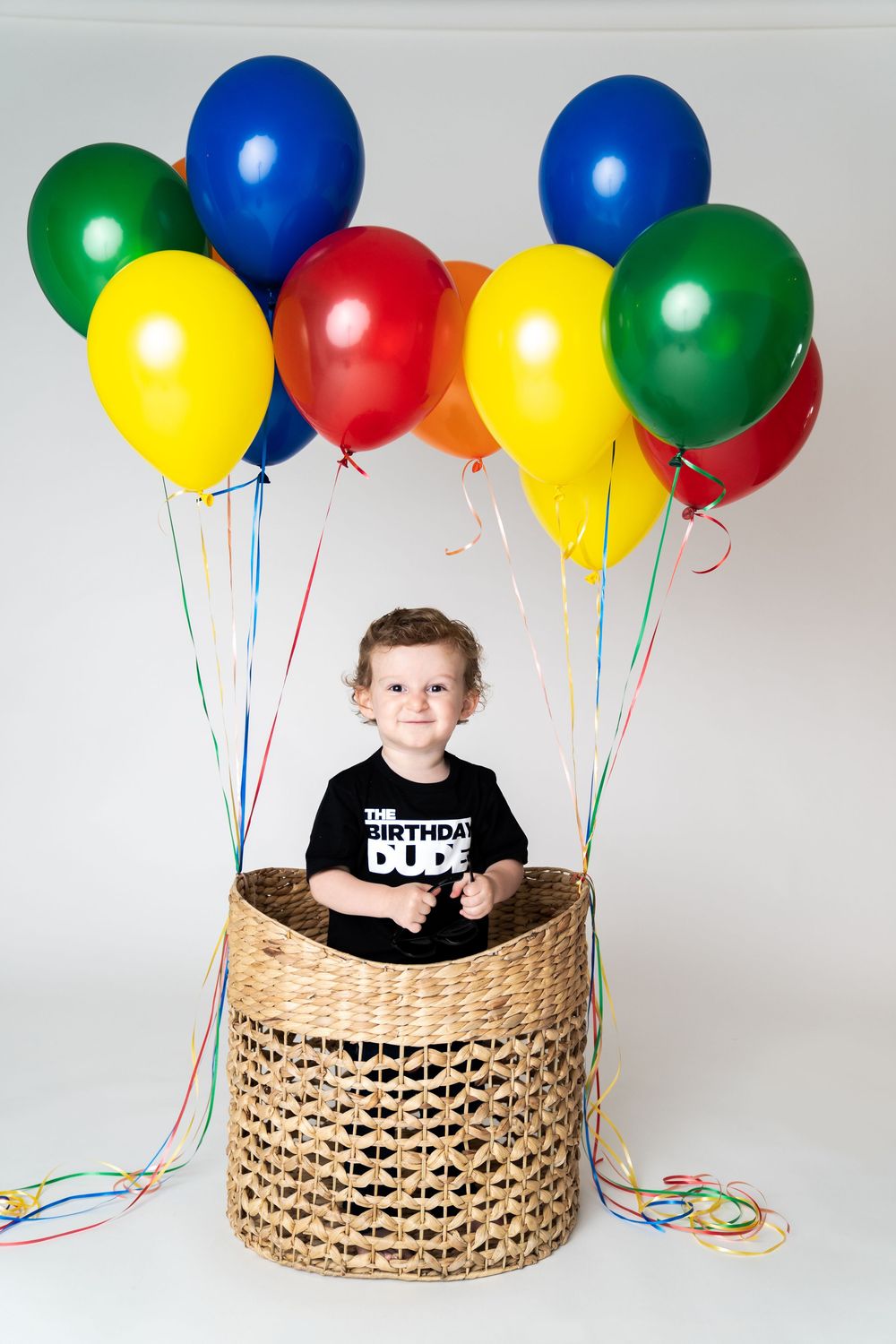Happy toddler boy in a black Birthday Dude shirt smiles inside a wicker basket with colorful balloons.