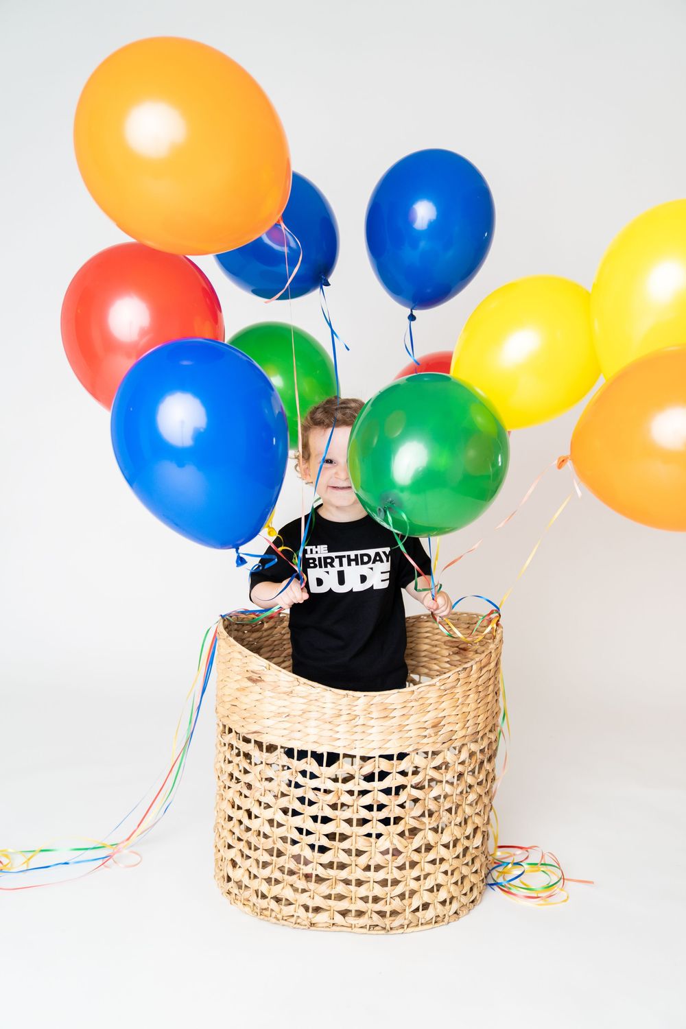 Young boy sitting in wicker basket surrounded by colorful balloons wearing a Birthday Dude shirt on white background.