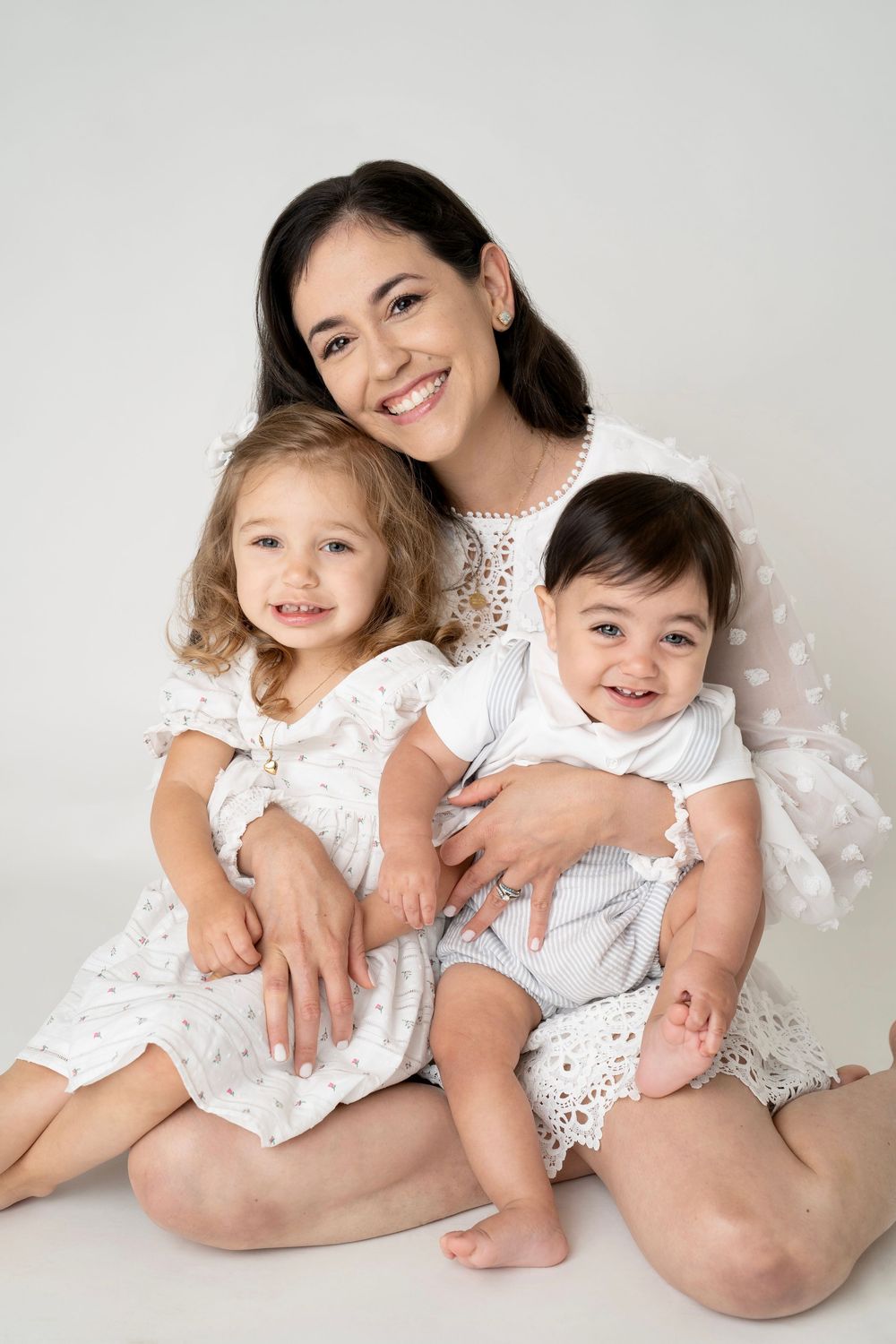 A smiling mother in white lace dress poses with her two young children, a toddler girl and baby boy, on white background.