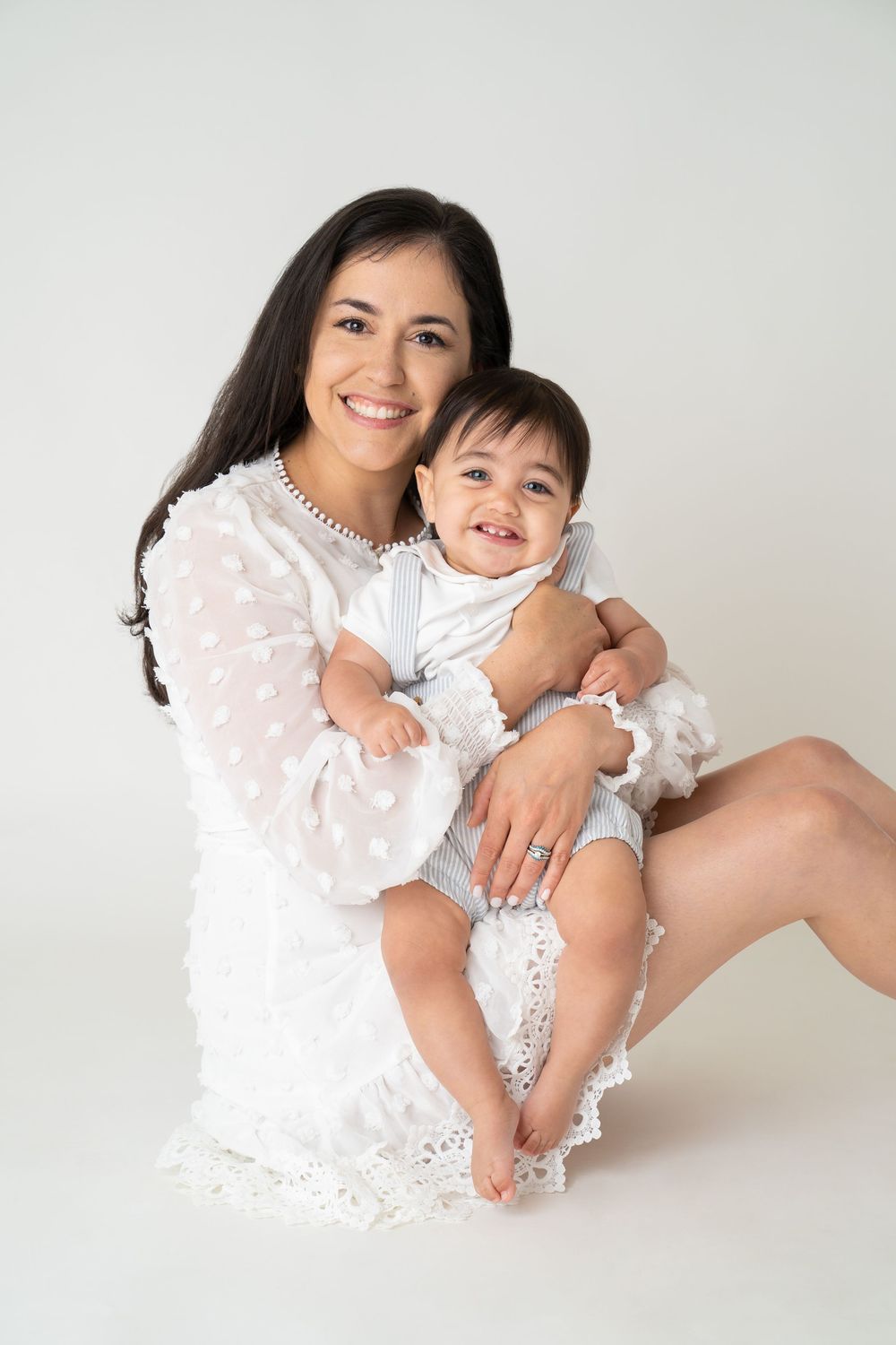 Smiling mother in white dress holding happy baby in light blue outfit against white background.