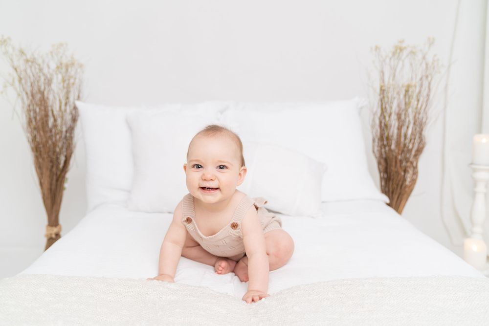 Smiling baby in beige onesie sitting on white bed with pillows and dried floral arrangements.