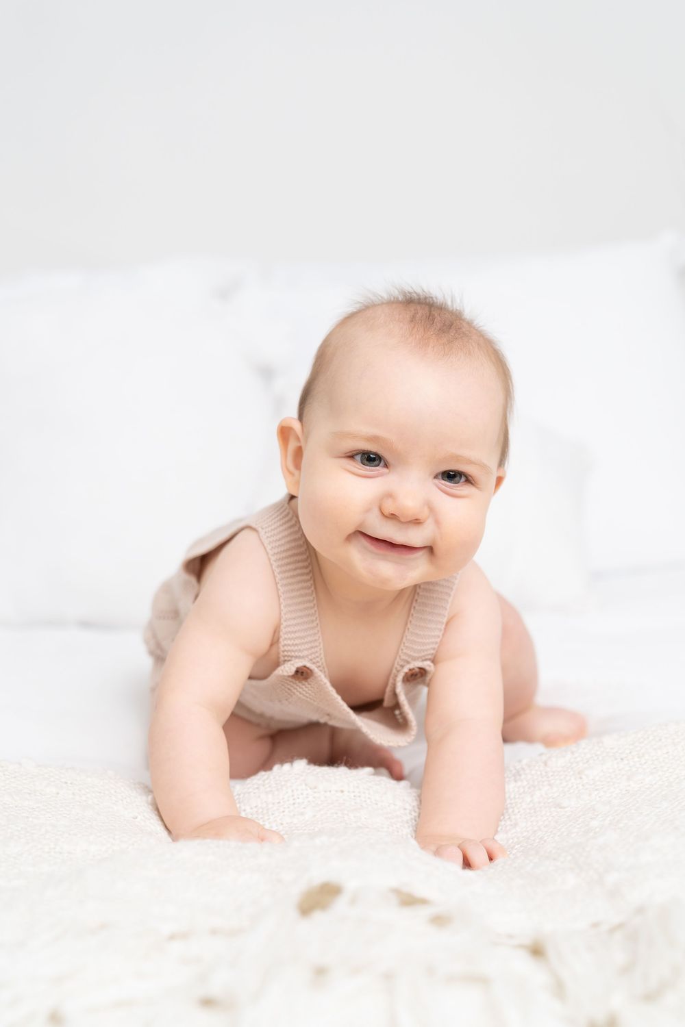 Smiling baby in beige romper doing tummy time on white fluffy blanket with pillows in background.
