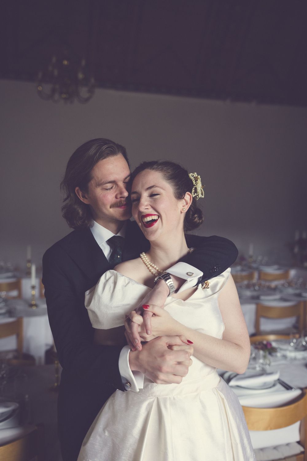Groom embracing laughing bride in vintage white dress inside decorated wedding reception venue.