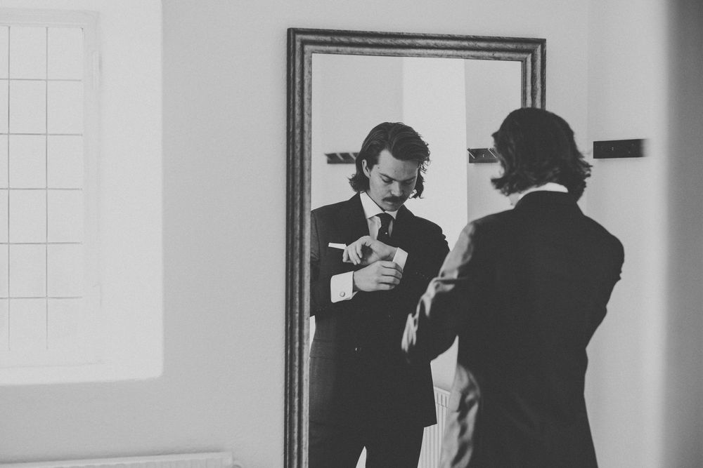 Black and white photo of groom adjusting cufflinks while checking his reflection in a framed mirror in Gothenburg