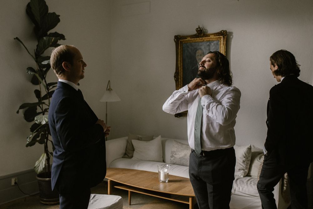 Groom adjusts tie while two men watch in a softly lit modern living room.
