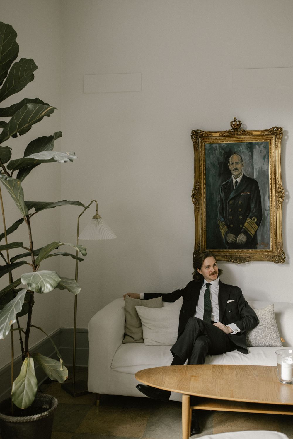 Groom in black suit sitting on white sofa beneath ornate gold-framed naval officer portrait with indoor plant nearby.