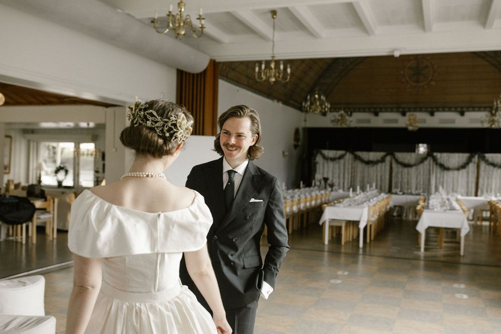Smiling groom in dark suit holding brides hand in elegant wedding reception hall with chandelier.