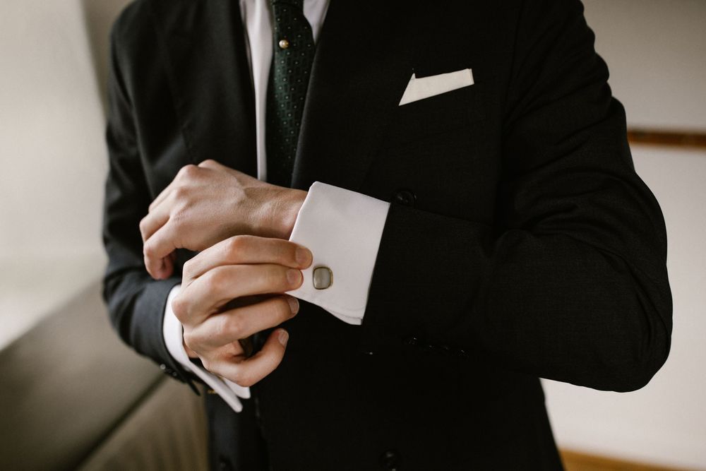 Close-up of a groom in a black suit fastening his silver cufflinks before the wedding ceremony.