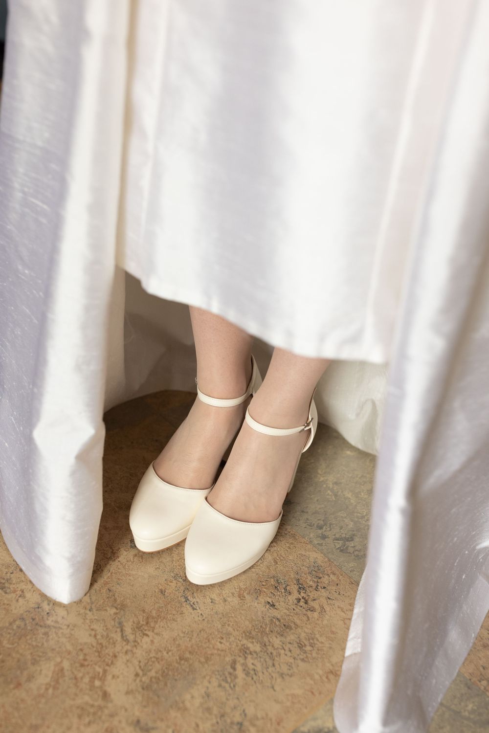 Bride's ivory block heel shoes with ankle strap peeking beneath a white satin wedding gown.