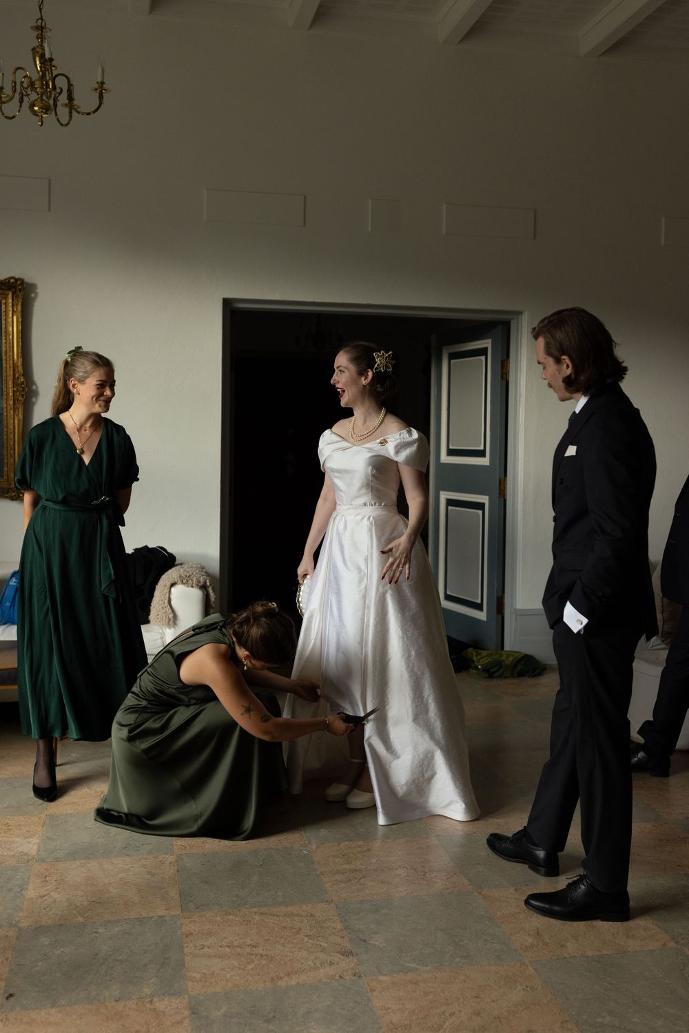 Bridesmaids in green dresses help laughing bride with wedding gown in elegant manor house.