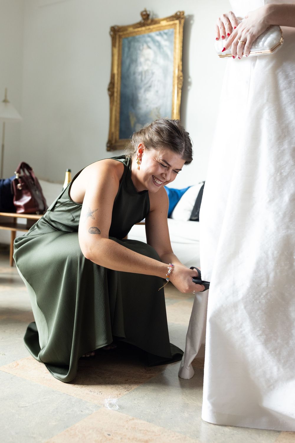 Smiling bridesmaid in olive green dress kneels to adjust bride's white wedding gown hem.