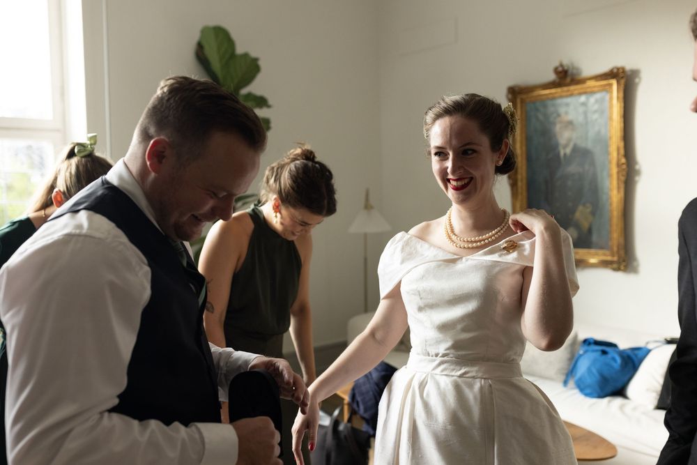 Bride in vintage white dress laughing with guest during pre-wedding preparations indoors.