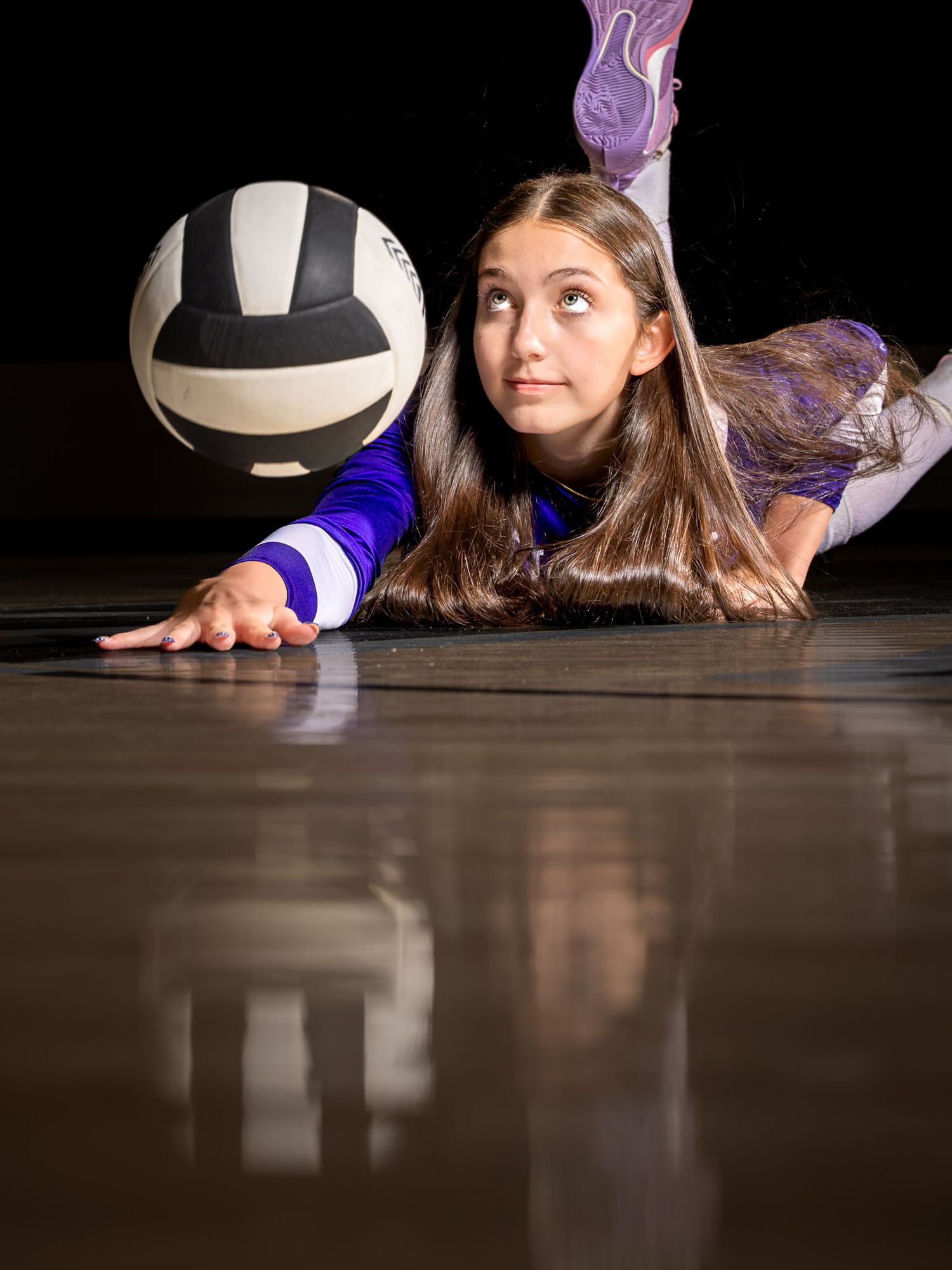 Young girl in purple volleyball uniform dives on court floor reaching for a black and white volleyball.