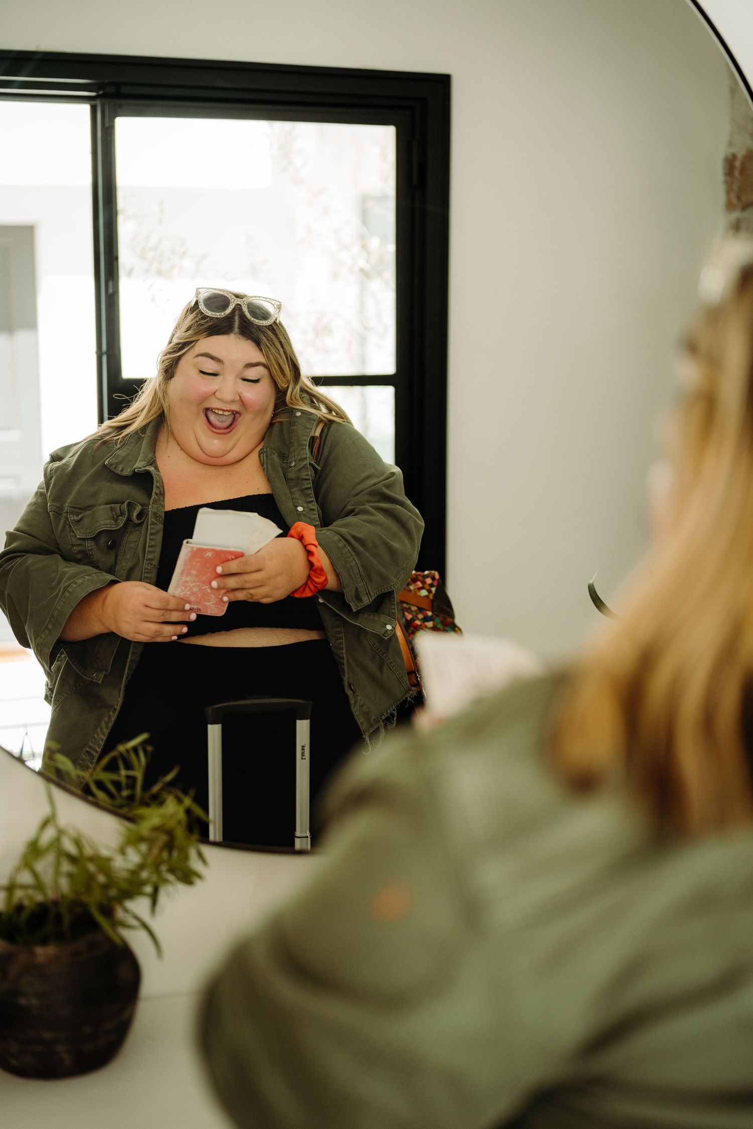 A plus-size woman in a green jacket laughs joyfully while holding cards, reflected in a modern mirror.