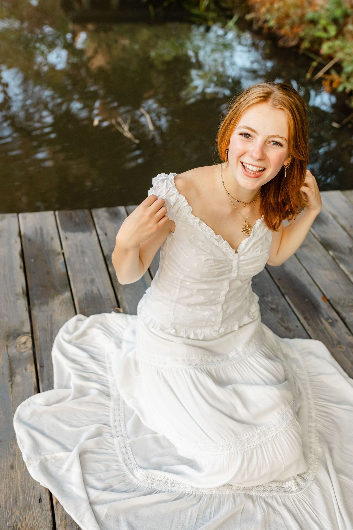 Smiling redhead bride in white wedding dress sitting on wooden dock by water in sunny outdoor setting.