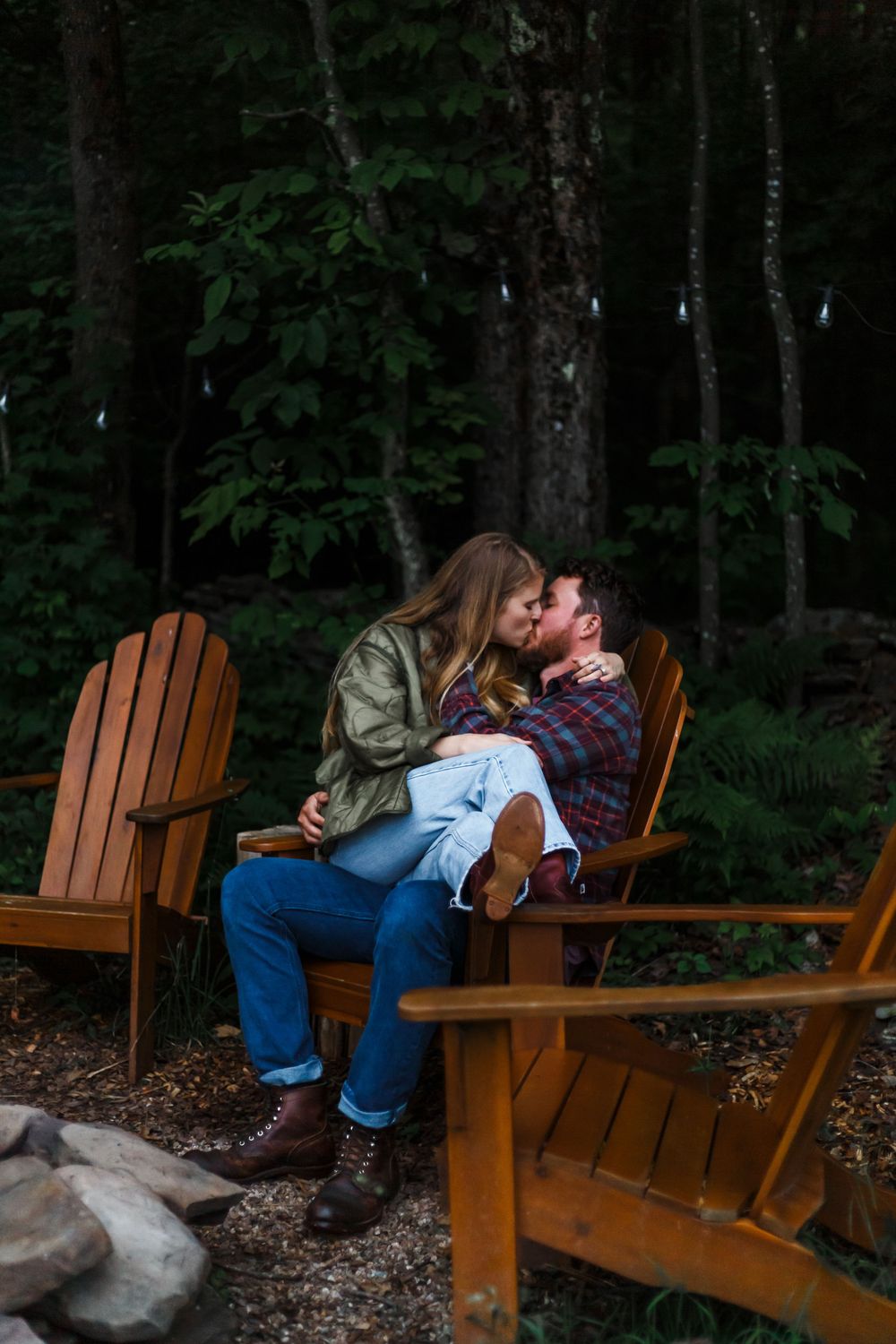 Couple kissing on Adirondack chairs by a fire pit surrounded by lush green forest trees.