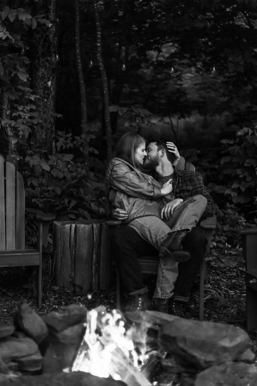 Black and white photo of a romantic couple kissing by a campfire in a forest setting.
