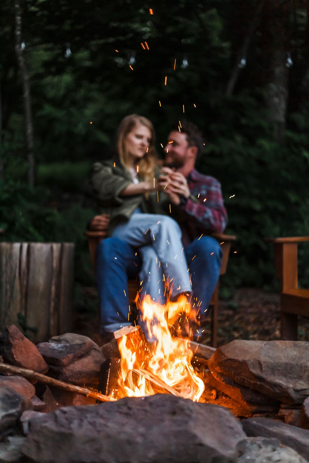 Couple sitting in wooden chairs by a crackling campfire with glowing sparks in a forest setting.