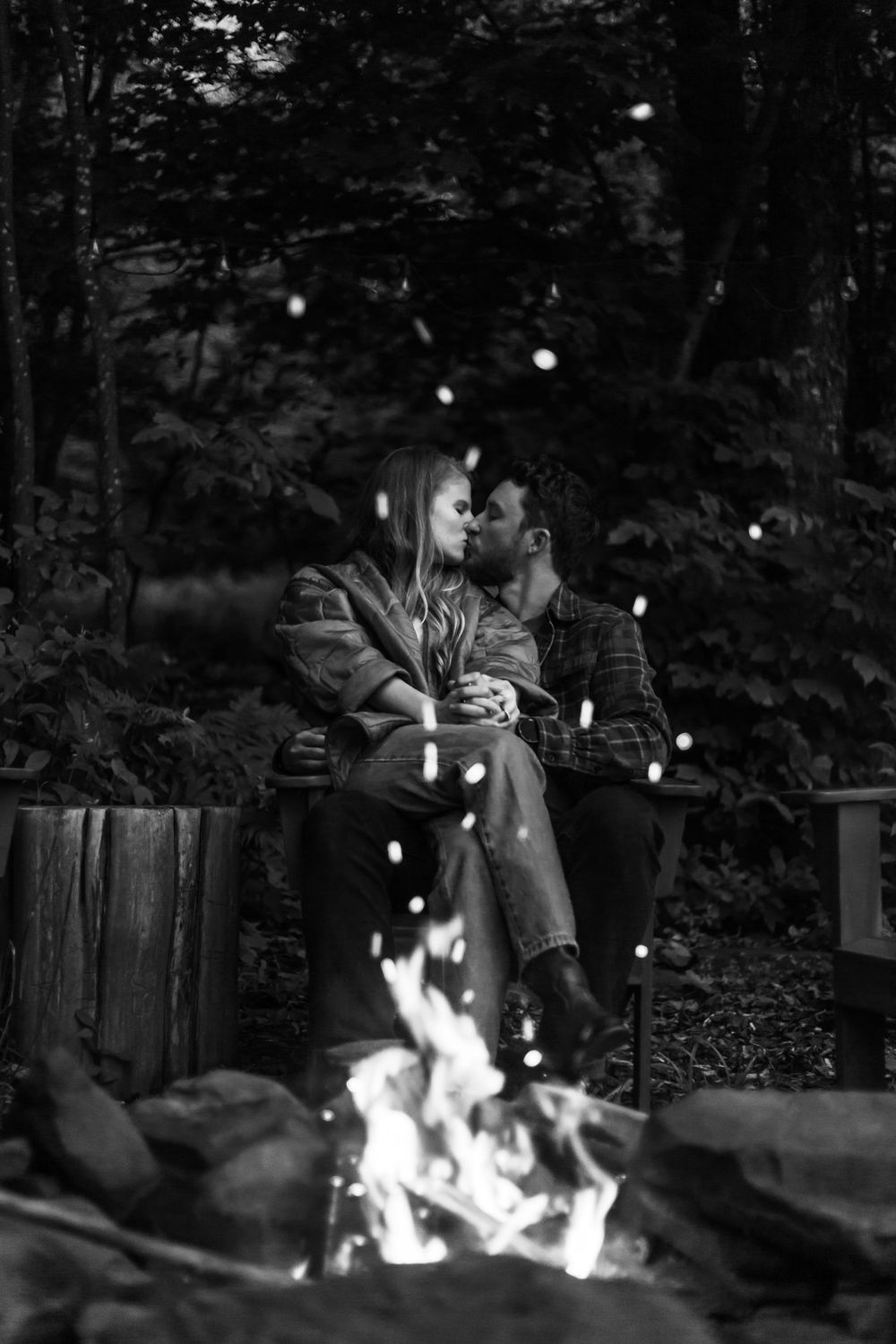 Black and white photo of a romantic couple kissing by a glowing campfire in a forest at night.