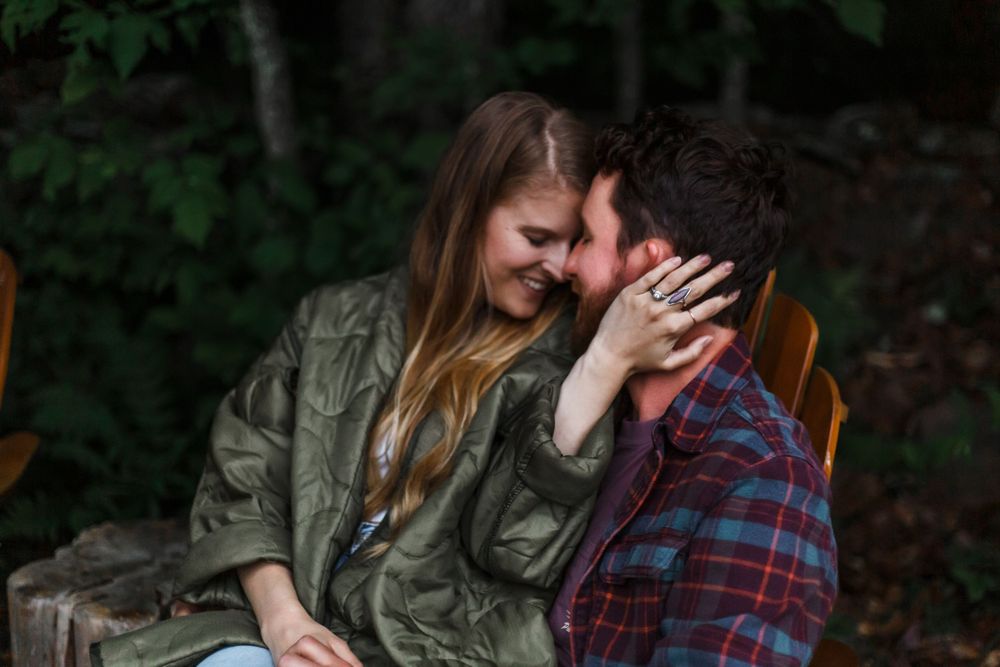 A couple shares a romantic moment outdoors, smiling closely together, woman in green jacket, man in plaid shirt.