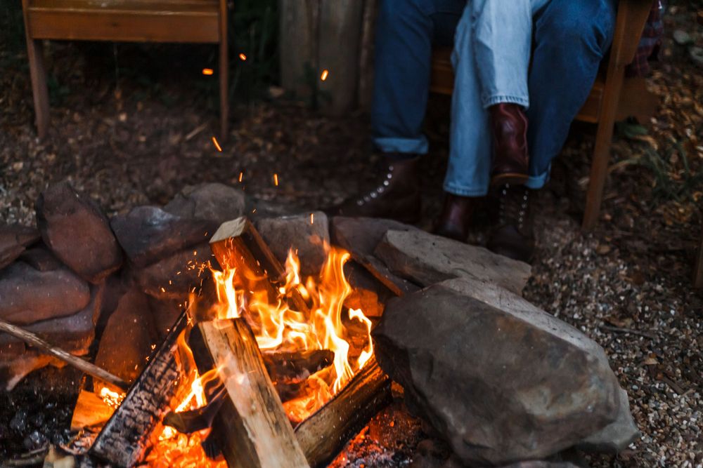 Person relaxing by a rustic campfire surrounded by rocks, with glowing flames and wood logs burning outdoors.