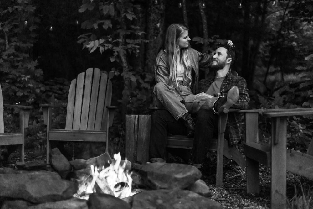 Couple sitting by a campfire in Adirondack chairs surrounded by trees in a black and white photo.