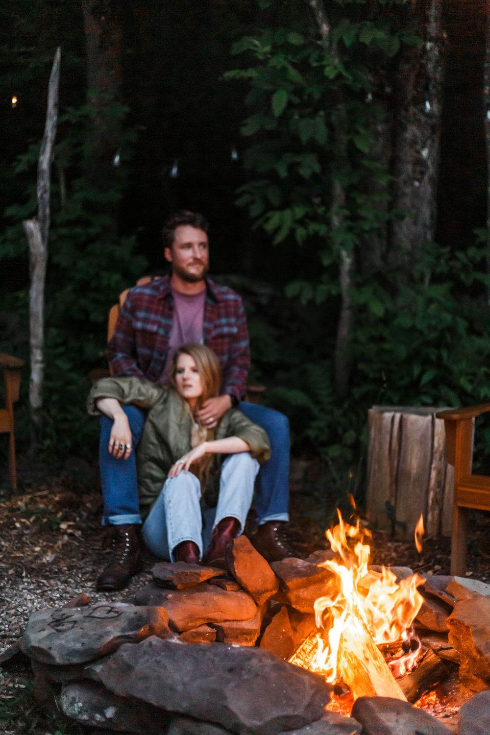 Couple sitting together by a campfire in a forest at dusk, woman in green jacket leaning against man in plaid shirt.
