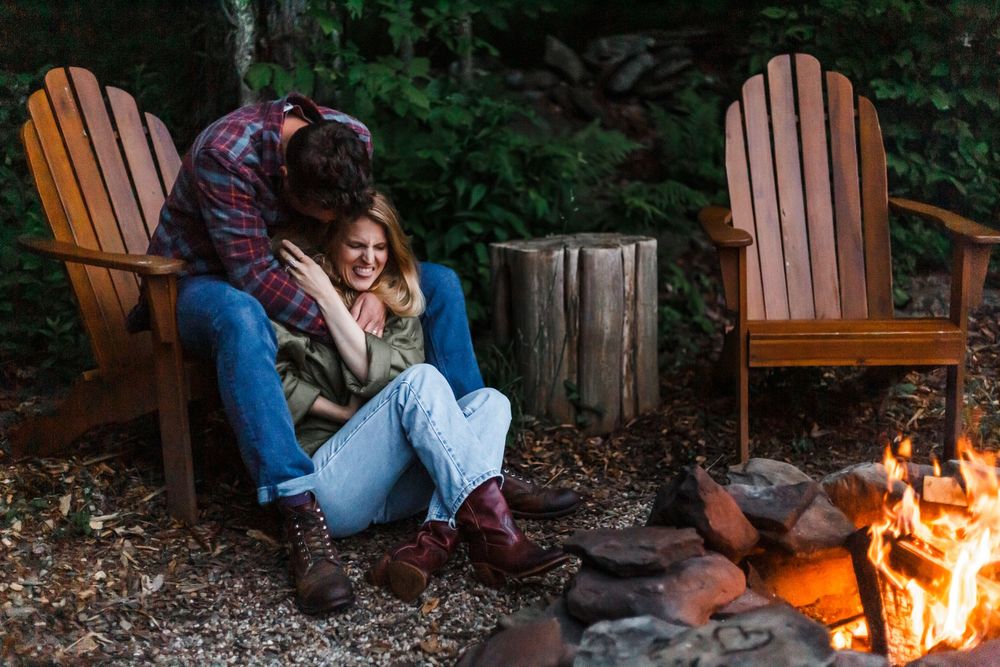 Romantic couple cuddling together in Adirondack chairs beside a warm campfire in the woods.
