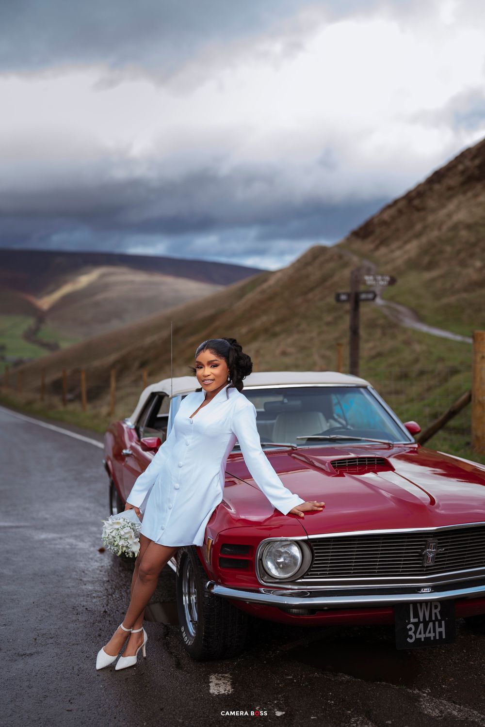 Bride in white dress poses by a classic red Ford Mustang on a scenic road with rolling hills.