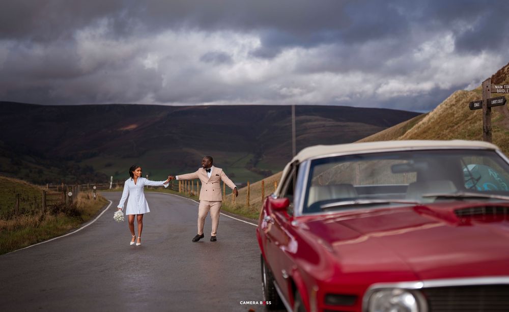 Couple holding hands on a scenic road with rolling hills, dramatic clouds, and a classic red car in the foreground.
