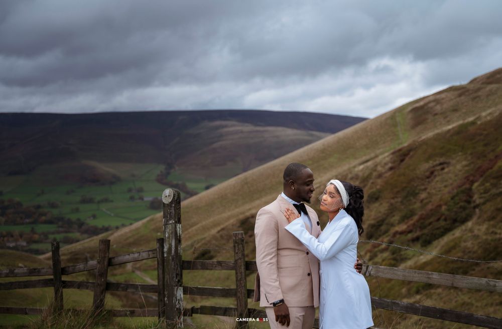 Couple sharing a romantic moment by a wooden fence with rolling green hills and dramatic cloudy skies.