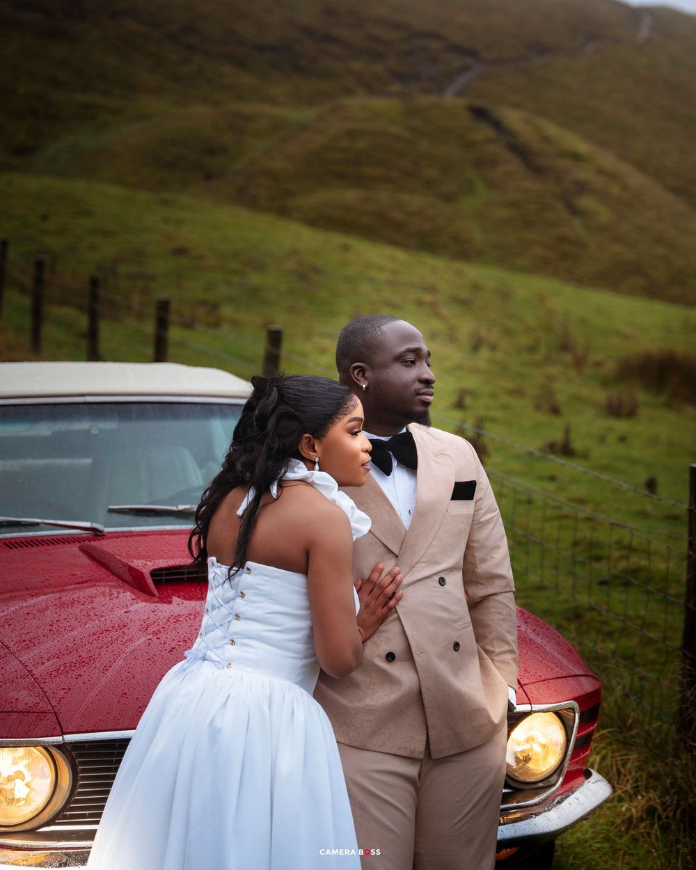 Couple in wedding attire posing by a vintage red car with green hills in the background.