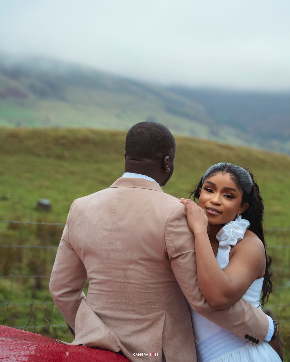 A couple in formal attire poses romantically against a red surface with lush green hills in the background.