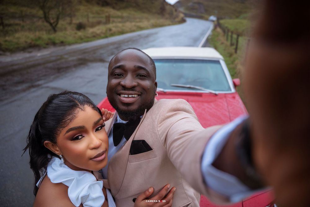 Couple taking a selfie in front of a red classic car on a scenic road, dressed in elegant formal attire.
