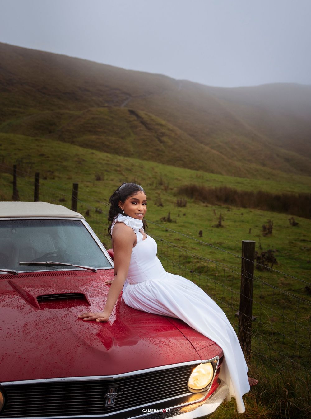 Woman in white dress posed on red classic car hood with misty green hills in background.