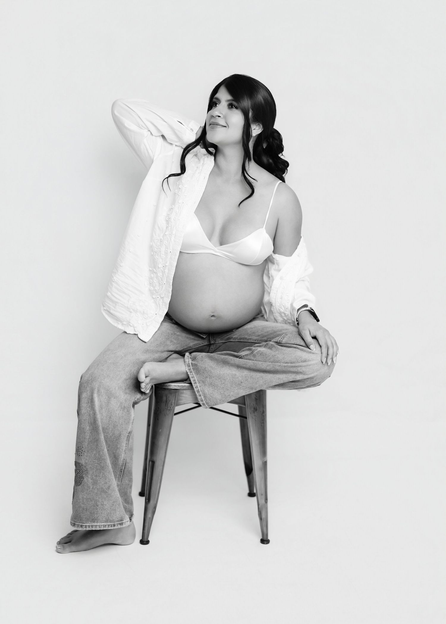 Black and white maternity photo of a pregnant woman sitting on a chair in a bra top, open shirt, and jeans.