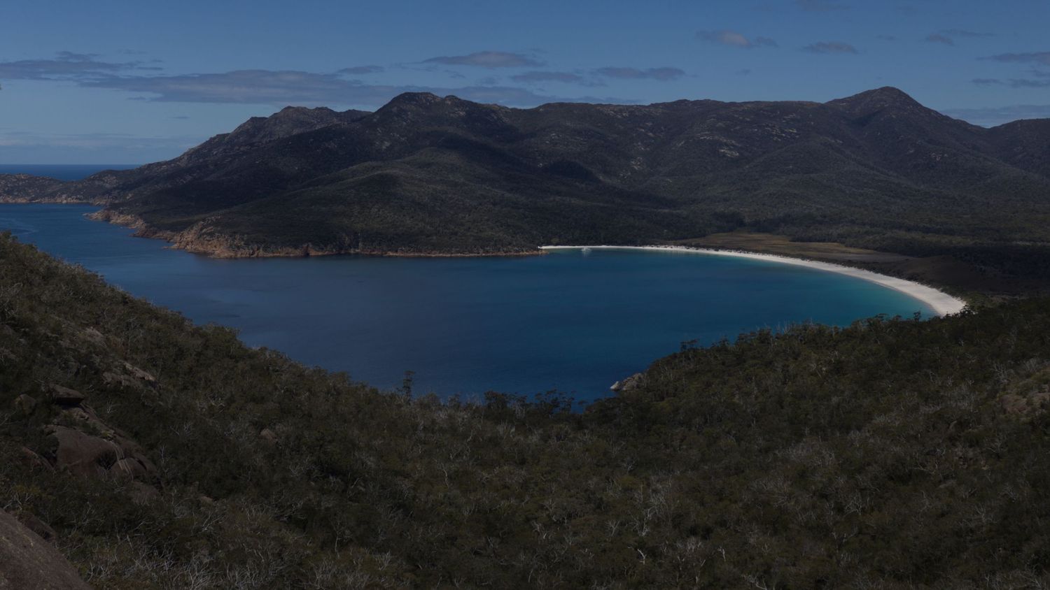 Aerial view of Wineglass Bay, Tasmania, with turquoise waters, white sandy beach, and lush green mountains.