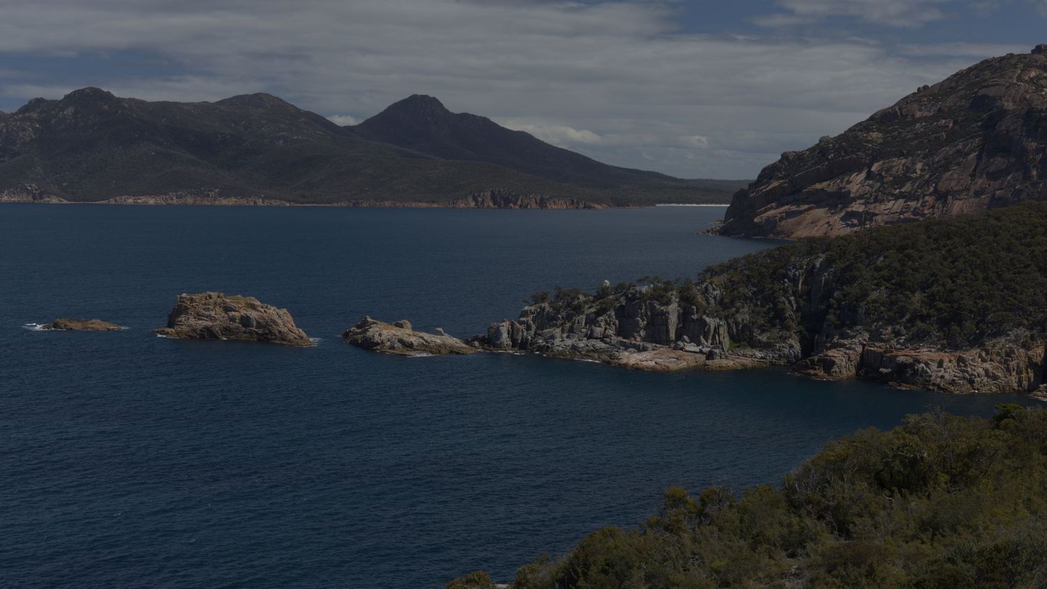 Rocky coastal landscape with blue ocean waters, rugged cliffs, and mountain peaks under a partly cloudy sky.