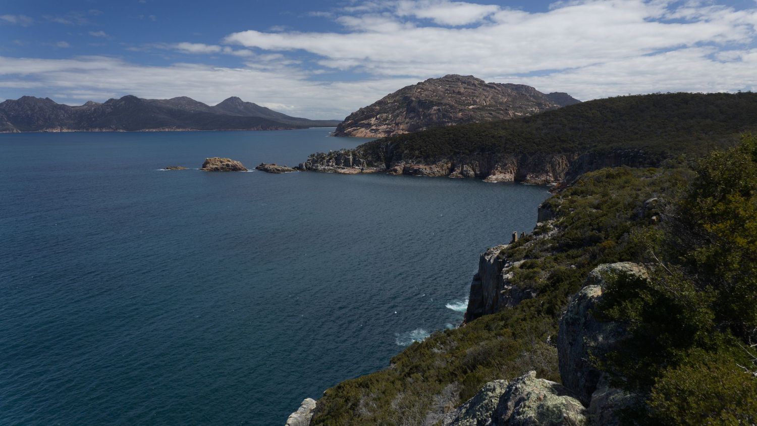 Aerial view of a stunning coastal landscape with turquoise waters, rocky cliffs, and lush green hills under a blue sky.