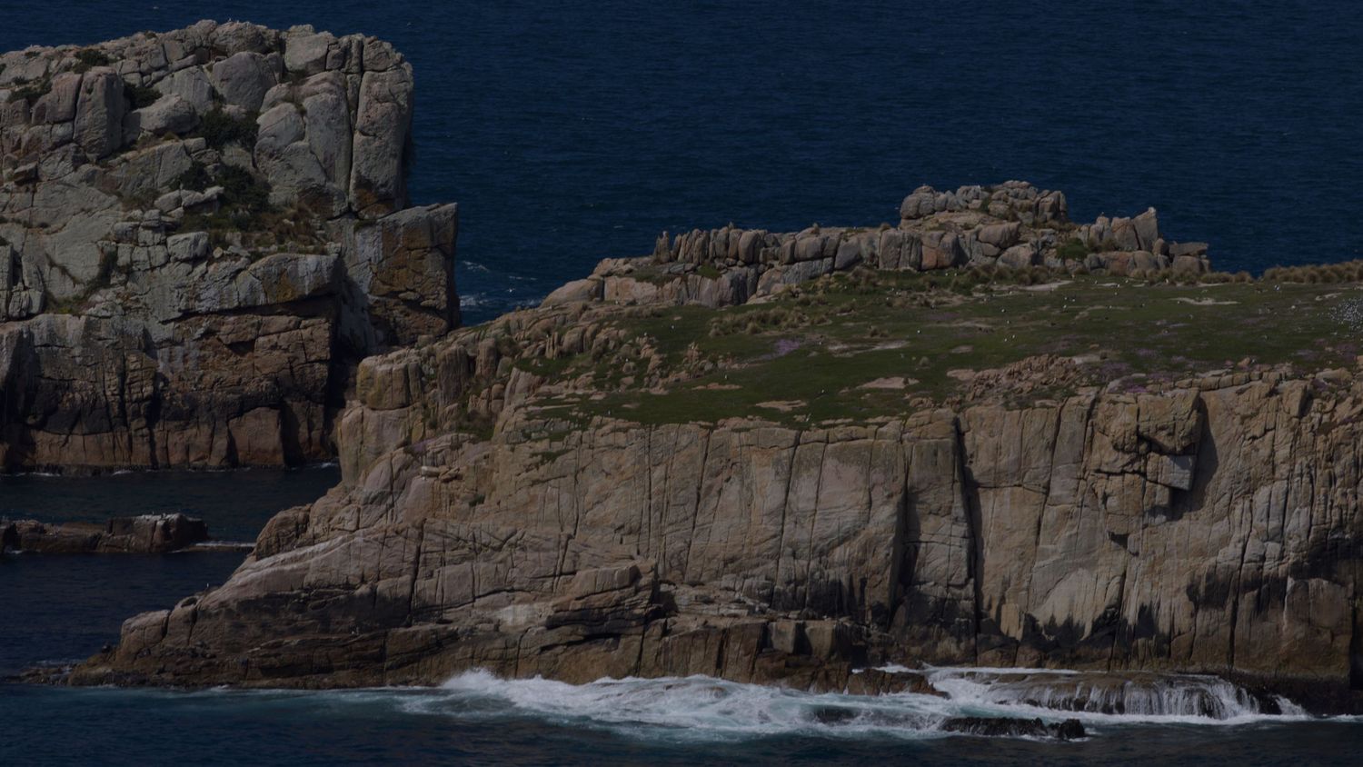 Rocky coastal cliffs with crashing waves below and green vegetation on top under a clear sky.