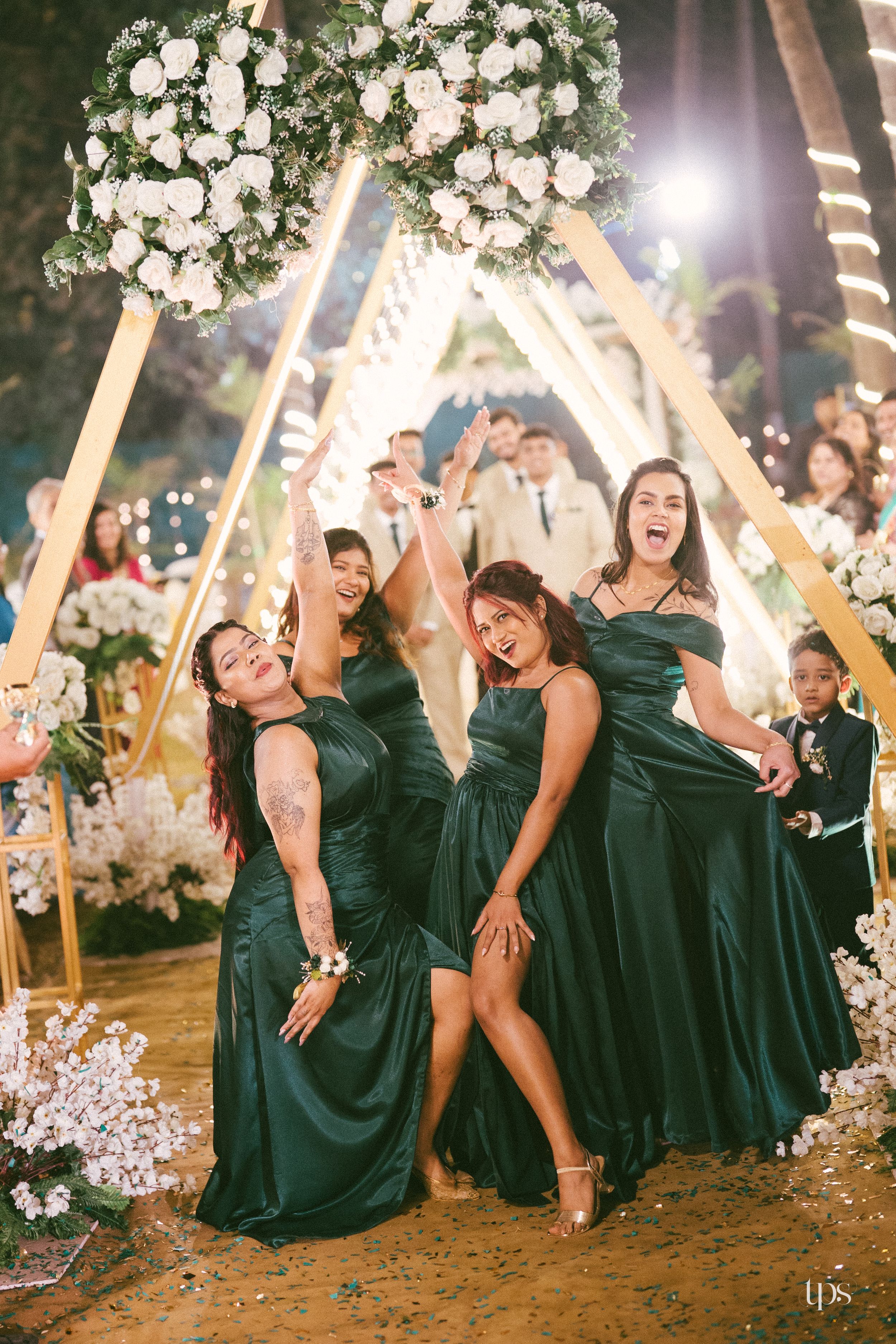 Bridesmaids in emerald green satin gowns celebrate joyfully under a floral arch at a wedding reception.