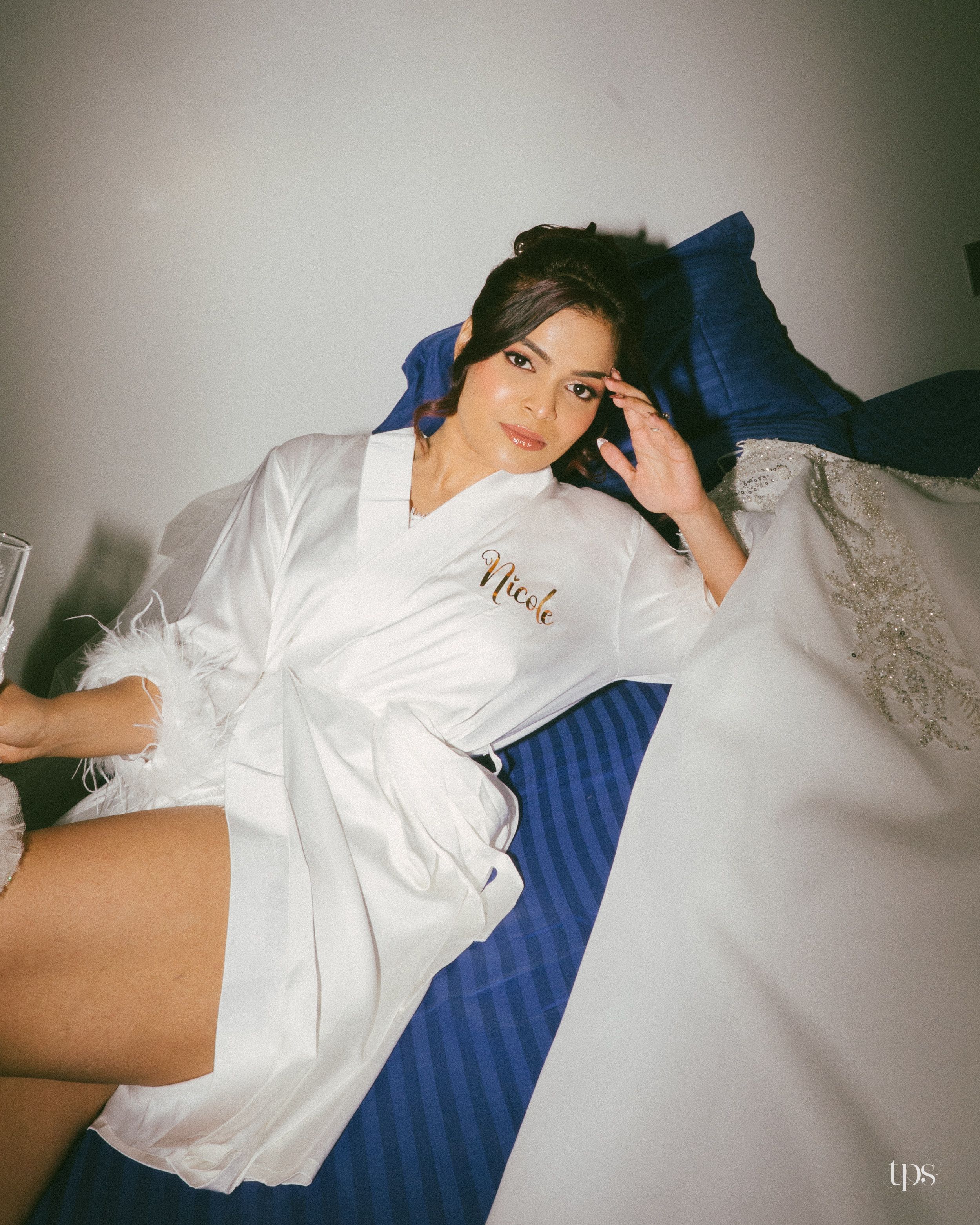 Bride in white satin robe with name embroidery relaxing on blue striped bed with lace wedding dress nearby.