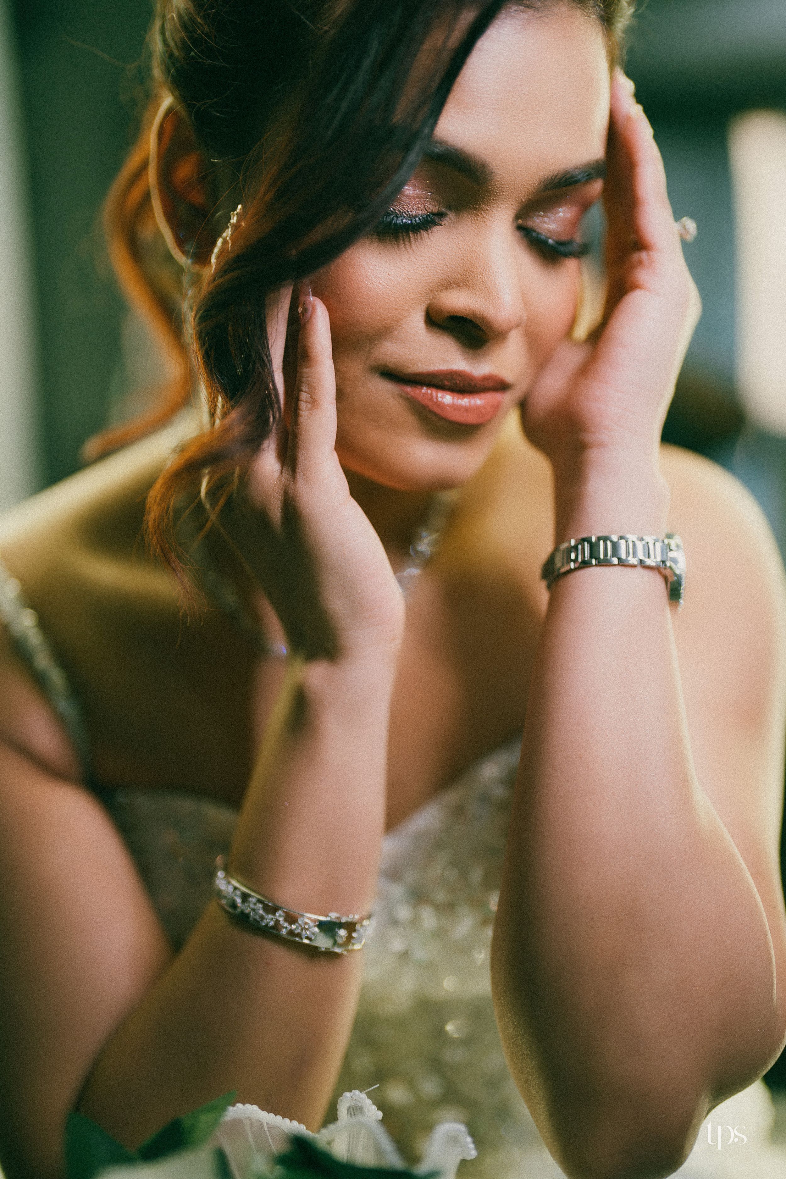Elegant bride with closed eyes, hands framing her face, wearing a white gown, diamond bracelets, and a silver watch.