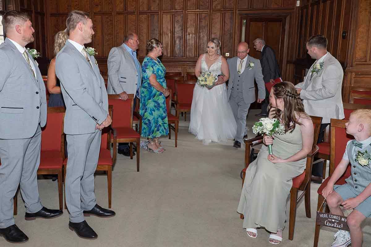 Bride and groom walking down the aisle in a wood-paneled wedding ceremony hall with guests and flower girl watching.